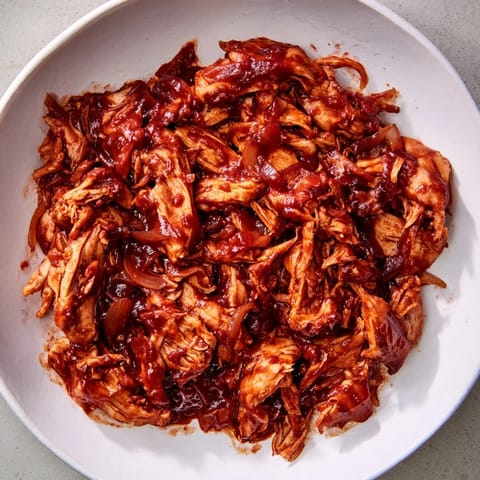 A close-up of tender, shredded BBQ pulled chicken, glistening with a rich, smoky barbecue sauce.